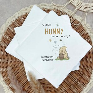 Puede incluir: Servilletas de papel blanco con una ilustraci&oacute;n impresa de un oso marr&oacute;n claro sosteniendo un tarro de miel. El texto dice A little HUNNY is on the way! BABY NATHAN MAY 2, 202X, acompa&ntilde;ado de peque&ntilde;as mariposas coloridas y una trayectoria punteada.