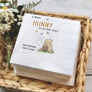 Puede incluir: Una pila de servilletas de papel cuadradas blancas con una ilustraci&oacute;n impresa de un oso sentado con un tarro de miel. El texto dice A little HUNNY is on the way! junto con el nombre BABY NATHAN y la fecha MAY 2, 202X en letras negras.