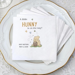 Puede incluir: Una pila de servilletas de papel blancas con una ilustraci&oacute;n impresa de un oso marr&oacute;n claro sentado junto a un tarro de miel. El texto dice A little HUNNY is on the way! junto con el nombre BABY NATHAN y la fecha MAY 2, 202X en tinta negra.