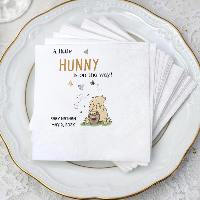 Puede incluir: Una pila de servilletas de papel blancas con una ilustraci&oacute;n impresa de un oso marr&oacute;n claro sentado junto a un tarro de miel. El texto dice A little HUNNY is on the way! junto con el nombre BABY NATHAN y la fecha MAY 2, 202X en tinta negra.