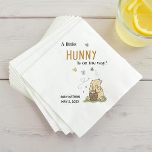 Puede incluir: Una pila de servilletas de papel blancas con una ilustraci&oacute;n impresa de un oso sentado junto a un tarro de miel. El texto dice A little HUNNY is on the way! junto con BABY NATHAN y MAY 2, 202X. Tambi&eacute;n hay mariposas y una abeja impresas.