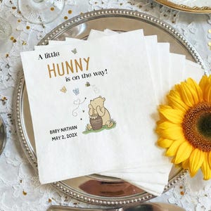 Puede incluir: Una pila de servilletas de papel blancas con una ilustraci&oacute;n impresa de un oso sosteniendo un tarro de miel, rodeado de peque&ntilde;as mariposas y abejas. El texto dice A little HUNNY is on the way! BABY NATHAN MAY 2, 202X en letras negras y doradas.