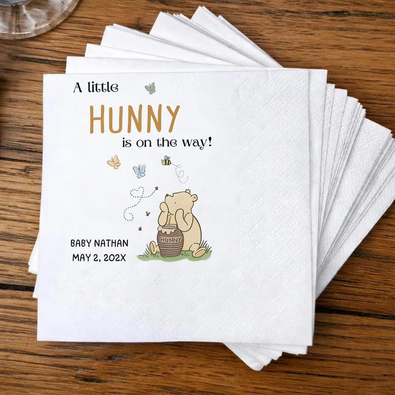 Puede incluir: Una pila de servilletas de papel blancas con una ilustraci&oacute;n impresa de un oso marr&oacute;n claro que sostiene un tarro de miel con la etiqueta HUNNY. El texto dice A little HUNNY is on the way! BABY NATHAN MAY 2, 202X, junto a mariposas y una abeja.