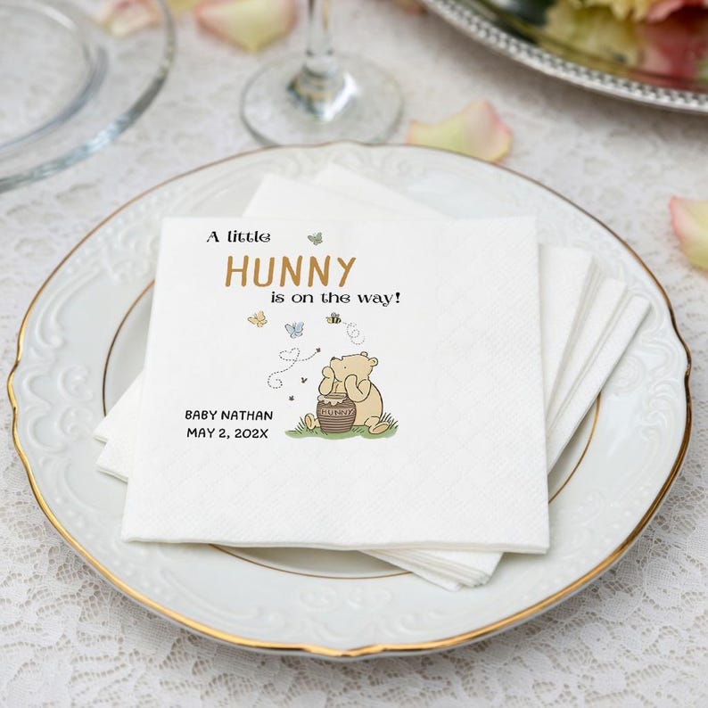 Puede incluir: Una servilleta de papel cuadrada blanca con una ilustraci&oacute;n impresa de un oso sosteniendo un tarro de miel, rodeado de peque&ntilde;as mariposas y abejas. El texto dice A little HUNNY is on the way! BABY NATHAN MAY 2, 202X en letras negras y doradas.