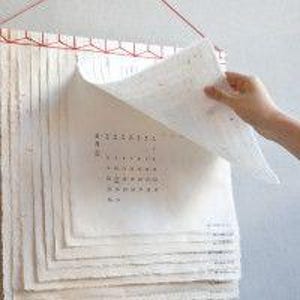 以下が含まれることがあります： 手作りの紙の束で、そのうちの1枚にカレンダーが印刷されています。紙は赤い糸と木製のフレームでまとめられています。カレンダーはめくられて、次の月が表示されています。