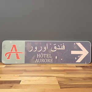 Könnte beinhalten: Ein blaues und weißes Schild mit einem Pfeil, der nach rechts zeigt. Das Schild zeigt "Fندق اورور" in Arabisch und "HÔTEL AURORE" in Französisch.
