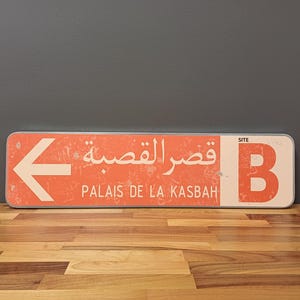 Könnte beinhalten: Ein rot-weißes Schild mit einem nach links zeigenden Pfeil. Das Schild zeigt "Palais de la Kasbah" auf Französisch und "قصر القصبة" auf Arabisch. Das Schild hat auch ein weißes "B" auf rotem Hintergrund mit dem Wort "SITE" darüber.