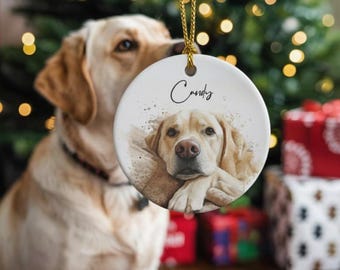May include: A round, white Christmas ornament with a watercolor image of a yellow Labrador Retriever. The dog's name, "Candy", is written in cursive above the image. A gold string is attached for hanging. The background includes a Christmas tree and wrapped gifts.