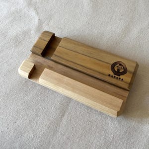 May include: A wooden phone stand with a light and dark wood grain. The stand has a slot for the phone and a raised section to hold the phone in place. The word "GABAKA" is printed on the stand.