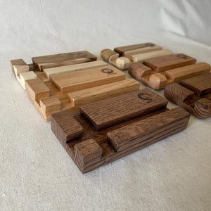 May include: A collection of wooden phone stands in various shades of brown. Each stand features a rectangular base with a slot for a phone and a small, engraved logo. The wood grain is visible, showcasing the natural texture.