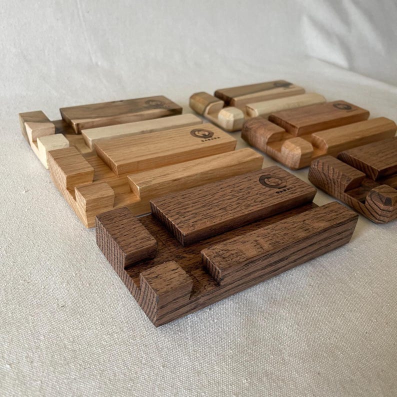 May include: A collection of wooden phone stands in various shades of brown. Each stand features a rectangular base with a slot for a phone and a small, engraved logo. The wood grain is visible, showcasing the natural texture.