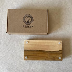 May include: A rectangular wooden tray with a natural wood grain pattern and four small white feet. The tray is next to a brown cardboard box with the logo "GABAKA HOME COMFORT" printed on it. The logo features a lightbulb inside a tree ring.