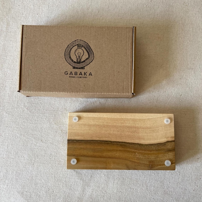 May include: A rectangular wooden tray with a natural wood grain pattern and four small white feet. The tray is next to a brown cardboard box with the logo "GABAKA HOME COMFORT" printed on it. The logo features a lightbulb inside a tree ring.