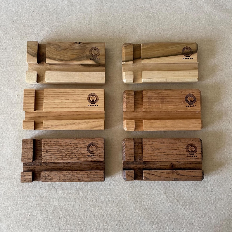 May include: Six wooden phone stands in various shades of brown, from light to dark. Each stand has a unique wood grain pattern and a slot for holding a phone. The stands are labeled with the word "BABAKA" and a circular logo.