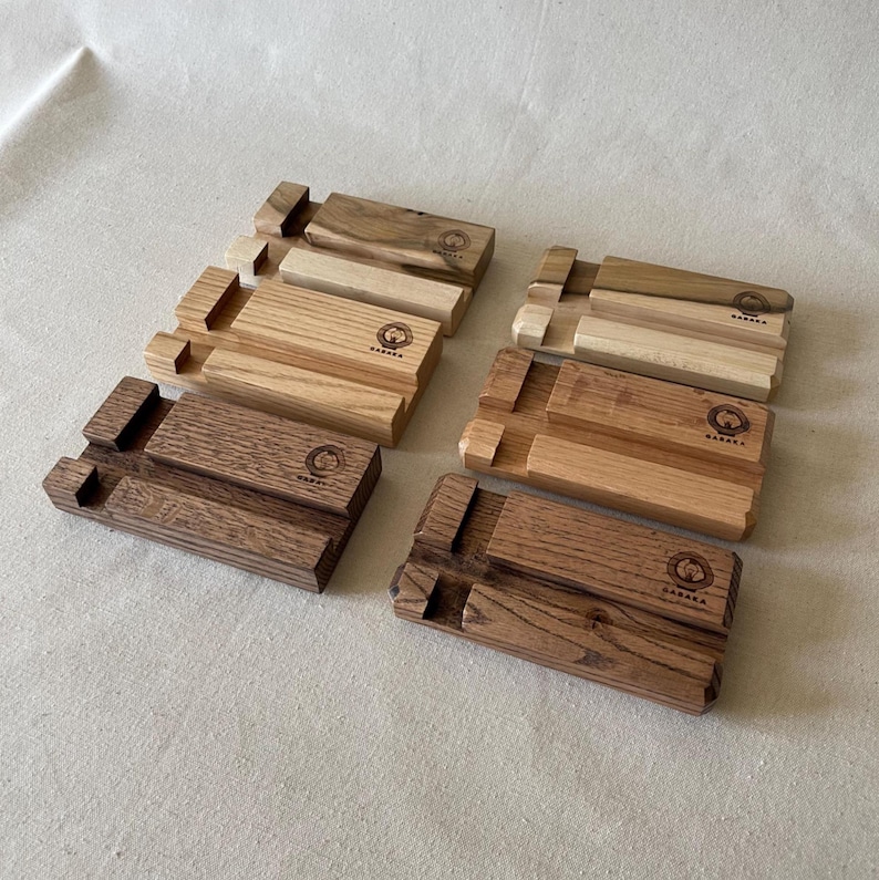 May include: A collection of wooden phone stands in various shades of brown and tan. Each stand features a slot for holding a phone and a logo. The stands are made of natural wood and have a simple, minimalist design.
