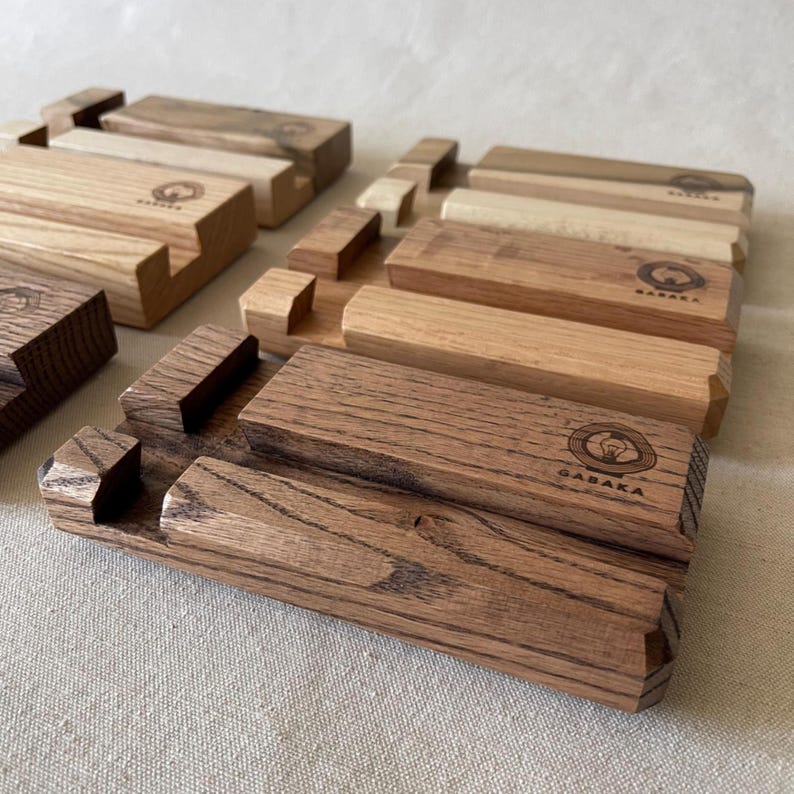 May include: Wooden phone stands in various shades of brown, with a light-colored surface. The stands have a slot for holding a phone and are engraved with the word "GABAKA" and a circular logo. The wood grain is visible.