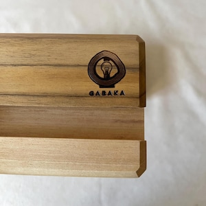 May include: A light-colored wooden phone stand with a recessed shelf. The stand features a laser-etched logo of a lightbulb inside a tree ring design, with the word "GABAKA" below. The wood grain is visible, and the stand has a clean, modern design.