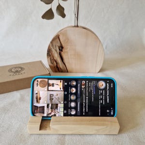 May include: A light-colored wooden phone stand with a slot to hold a phone. A blue-cased phone displays a social media page. A wooden vase and a cardboard box are in the background. The social media page shows the text "gabaka_home_comfort".