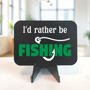 May include: Black rectangular sign with the text "I'd rather be FISHING" in white and green. The sign is supported by a black easel. The background is a blurred interior with plants.