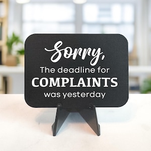 May include: A black sign with rounded corners, supported by a black stand. The sign features white text that reads "Sorry, The deadline for COMPLAINTS was yesterday."