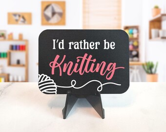 May include: A black sign with rounded corners, displaying the text "I'd rather be Knitting" in white and pink script. A white yarn ball and thread design is in the lower left corner. The sign is propped up on a black stand.