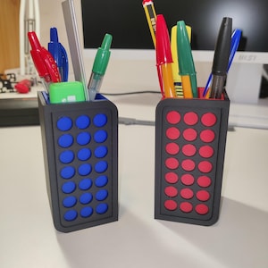 Puede incluir: Dos portalápices negros, uno con círculos azules y otro con círculos rojos, cada uno lleno de bolígrafos, lápices y una regla plateada. Los bolígrafos son de varios colores, incluyendo rojo, verde, azul y amarillo. Los soportes están sobre una superficie blanca.