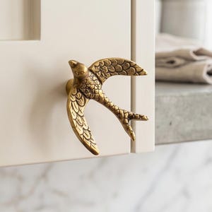 May include: A brass-toned metal cabinet knob shaped like a bird in flight with outstretched wings and detailed feather textures. The knob is mounted on a light-coloured cabinet door, featuring a metallic finish with visible dark antique-style accents.