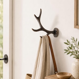 Könnte beinhalten: Ein an der Wand montierter Haken in Form eines dunkelbraunen Hirschgeweihs mit strukturierter Oberfläche. Der Haken ist mit einer runden Metallplatte an der Wand befestigt und hält einen beigen Stoffgegenstand mit einem braunen Lederriemen.