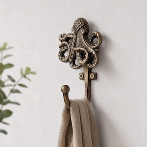 May include: A wall-mounted hook made of antique bronze-coloured metal, featuring a detailed octopus design at the top with curled tentacles. A single curved hook extends downward, holding a folded beige fabric cloth secured by two screws.