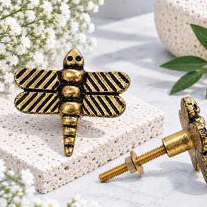 May include: Two dragonfly-shaped cabinet knobs made of antique gold-toned metal. Each knob features a detailed insect body with textured wings and a threaded metal bolt with a nut attached to the back for secure installation on furniture.