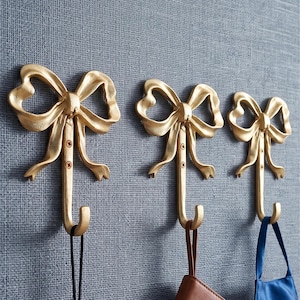 Puede incluir: Tres ganchos de pared de metal dorado, cada uno con forma de lazo decorativo con un gancho hacia abajo. Los ganchos están montados verticalmente y sostienen un estuche blanco de auriculares inalámbricos, un cuaderno marrón y una mascarilla azul.