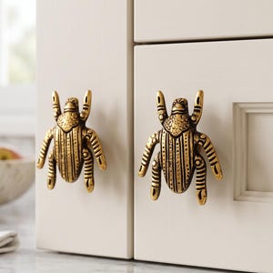 May include: Two gold-toned metal cabinet knobs shaped like beetles with detailed textured wings and legs. The knobs are mounted on light cream-colored cabinet doors, featuring a metallic finish with dark recessed areas to highlight the insect design.