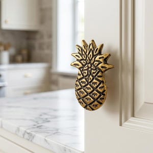 May include: A metallic gold-toned cabinet knob shaped like a pineapple with detailed textured leaves and a cross-hatched body. The hardware is mounted on a light-colored cabinet door, featuring a dark patina finish within the recessed grooves.