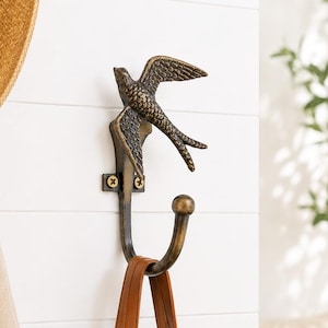 Pode incluir: Um gancho de parede feito de metal em tom de bronze escuro, com um design de andorinha esculpida no topo e um gancho curvo na parte inferior. A peça é fixada à parede com dois parafusos visíveis, proporcionando uma solução de armazenamento.