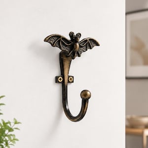 Puede incluir: Un gancho de metal montado en la pared que presenta una figura decorativa de murciélago con las alas extendidas en la parte superior. El gancho tiene un acabado de bronce antiguo con reflejos oscuros y se fija con dos tornillos visibles.