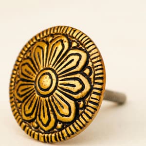May include: A decorative cabinet knob with a floral design. The knob is round, with a gold finish and black accents. The floral pattern is centered on the knob and surrounded by a textured border. The screw is silver.