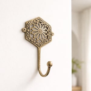 May include: Ornate brass wall hook with a hexagonal floral design. The hook has a decorative, curved shape and is mounted on a white wall. The hook is approximately 6 inches in length, perfect for hanging coats, hats, or other items.