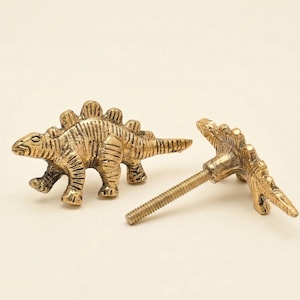 May include: Two gold-toned metal cabinet knobs shaped like Stegosaurus dinosaurs. Each knob features a textured, antique-style finish with visible plates along the back and a threaded metal screw extending from the rear for installation.