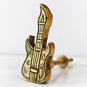 Puede incluir: Un pomo de mueble de metal en tono dorado con forma de guitarra eléctrica. La superficie presenta un acabado texturizado y desgastado con líneas negras grabadas que detallan el mástil, las cuerdas y patrones florales en el cuerpo. Un perno roscado sobresale por detrás.