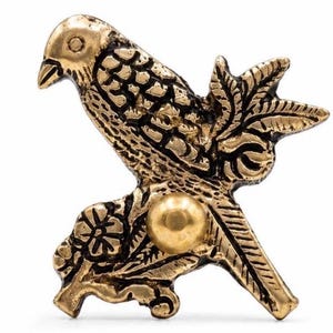 Puede incluir: Un broche detallado de pájaro dorado, posado en una rama con hojas y una flor. El pájaro tiene plumas texturizadas y un pequeño acento dorado redondo. El broche es probablemente de metal.