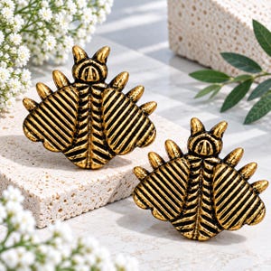 Peut inclure: Une paire d'objets décoratifs en métal doré en forme d'insecte, présentant un motif d'ailes nervurées détaillé et de petites pointes saillantes sur les bords. Les insectes ont un design symétrique avec un corps central et deux antennes.
