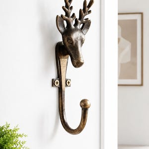 Könnte beinhalten: Ein an der Wand montierter Metallhaken mit einem skulptierten Hirschkopf an der Oberseite, detailliertem Geweih und einem geschwungenen Haken am unteren Ende. Das Stück hat ein dunkles, patiniertes Bronze-Finish und ist mit zwei sichtbaren Schrauben befestigt.