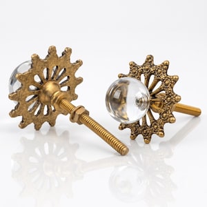 May include: Two cabinet knobs featuring a clear, spherical glass center set within an antique brass-toned metal base. The base has a sunburst or gear-like design with intricate, textured detailing and a threaded metal bolt for installation.