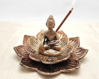 May include: A bronze Buddha statue sits on a lotus flower incense holder. The incense holder has a detailed petal design with a metallic finish. A lit incense stick emits a thin stream of smoke, adding a sense of tranquility.