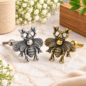 Peut inclure: Deux boutons de placard en métal en forme de scarabée, l'un avec une finition argentée et l'autre avec une finition dorée. Chaque bouton présente un design d'insecte détaillé avec des ailes texturées, un corps segmenté et une tige filetée.