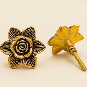 Puede incluir: Dos tiradores de armario antiguos con forma de flor de latón. Los tiradores tienen un diseño de rosa detallado en el centro y están montados en una varilla roscada con una tuerca. El acabado es un dorado cálido y envejecido.