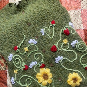 May include: A green knitted vest with embroidered flowers and strawberries. The flowers are yellow and white, and the strawberries are red.