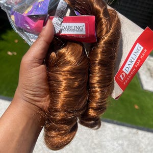 May include: A bundle of auburn-colored hair extensions, tightly curled, held in a clear plastic package. The product packaging includes a red label with the brand name "DARLING" and the tagline "Passion for Hair."