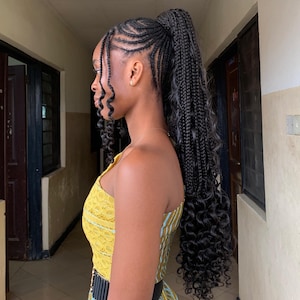 Puede incluir: Una mujer con cabello negro trenzado intrincadamente, peinado en una cola de caballo alta con extensiones largas y rizadas. Viste un vestido amarillo estampado con cinturón negro, de pie en un pasillo.