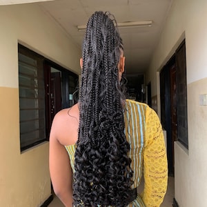 May include: A person with long, black braided hair styled in a high ponytail with loose, curly ends. The person is wearing a yellow and blue striped dress with a lace sleeve. The background is a hallway with doors and windows.